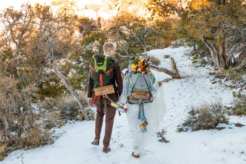 Bride and groom walking in winter snow at the grand canyon wearing just married backpacks for their elopement