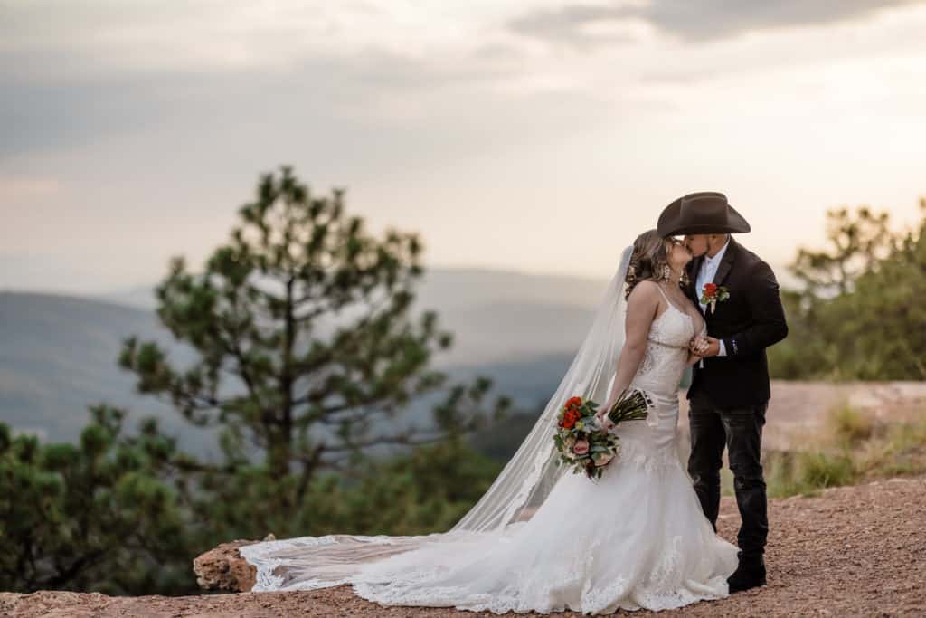 Bride and groom kissing at Mogollon Rim in Payson Arizona. Groom is wearing all black with a black cowboy hat. Bride's train is hanging off the edge of a cliff.
