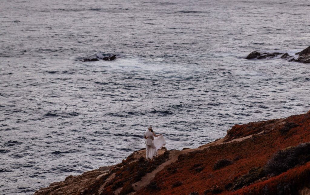 Bride holding wedding dress looking at groom. Standing on cliff in Big Sur, California during their sunset elopement.