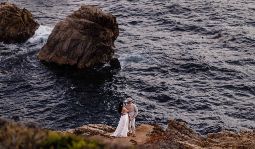 Bride and groom in wedding attire standing on rocky cliff holding each other in Big Sur, California.
