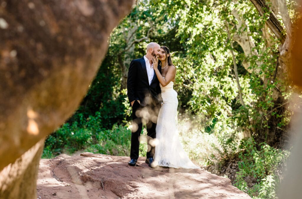 Boho bride holding her grooms face as he looks at her in front of oak creek in sedona during their intimate wedding